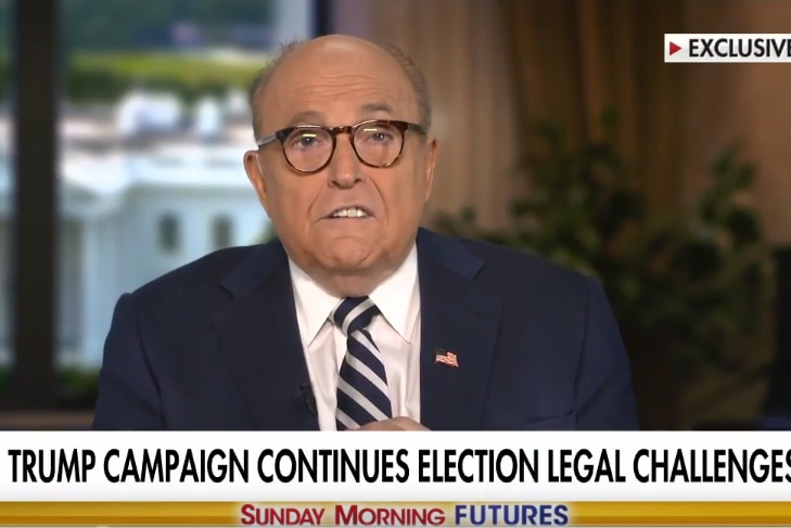 Rudy Giuliani speaking on a news television program wearing glasses and a suit and tie.