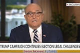 Rudy Giuliani speaking on a news television program wearing glasses and a suit and tie.