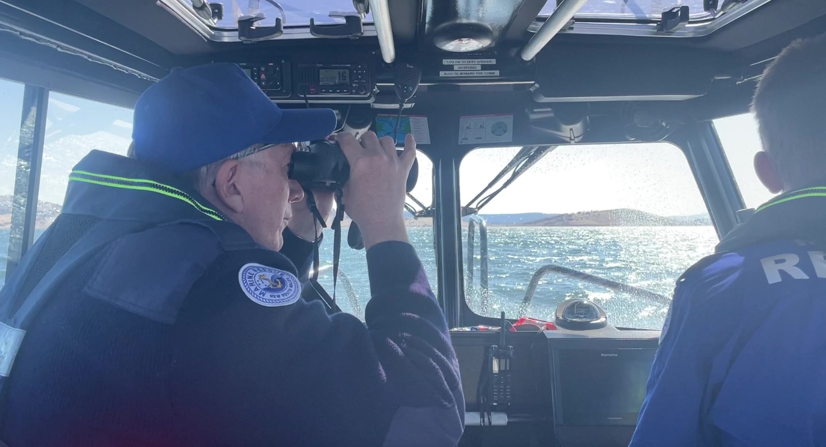 A man looks through binoculars over a lake, with another driving the boat