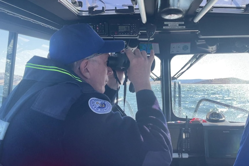 A man looks through binoculars over a lake, with another driving the boat