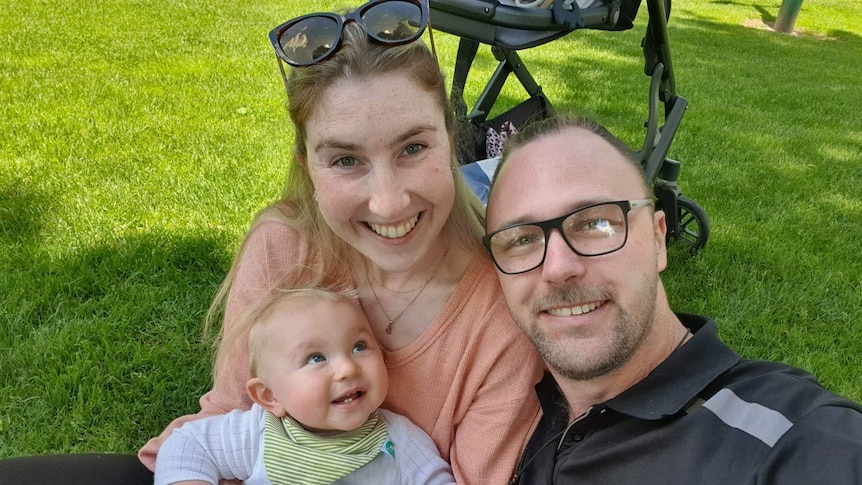 A woman and a man with a baby, all smiling as they sit together on some grass.