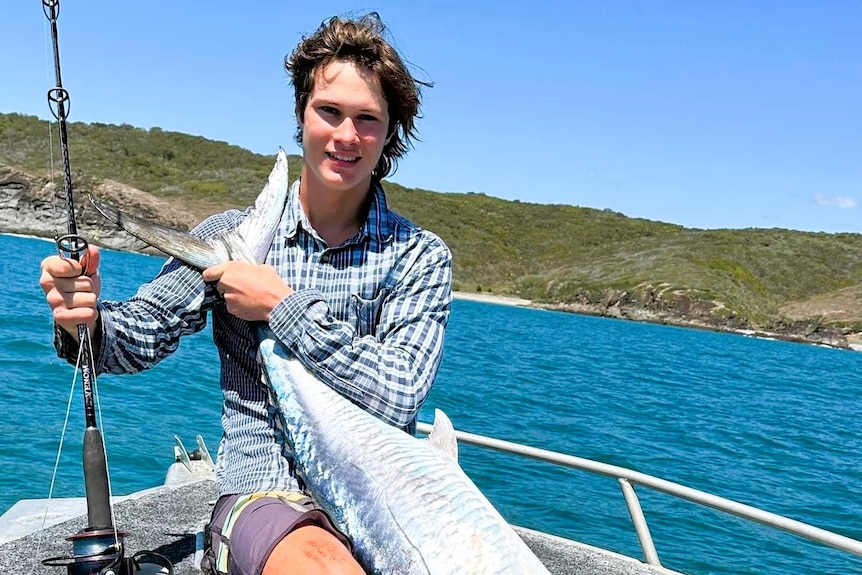 A teenage boy holding a fishing rod and Spanish mackerel fish on boat near an island and blue water.
