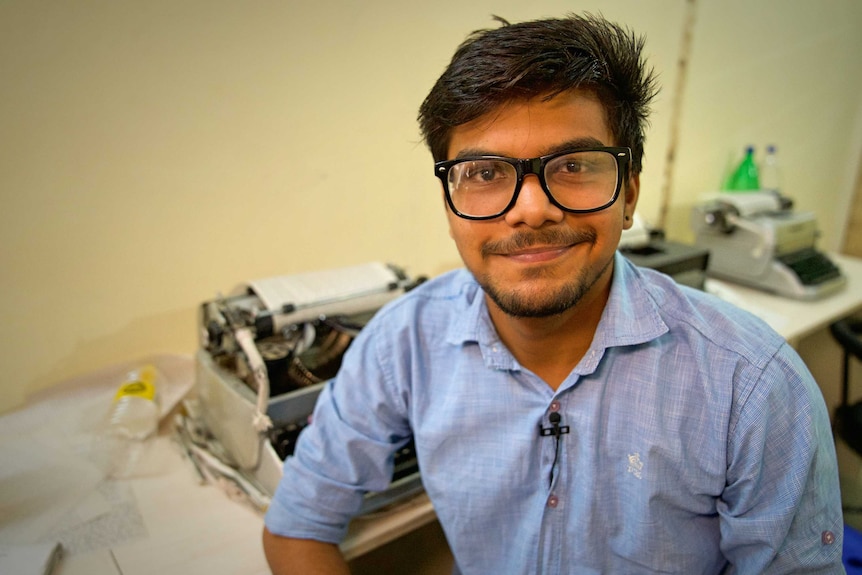 A young man in big glasses smiles as he sits at a desk with a typewriter