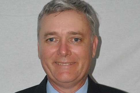 A man with grey hair and blue eyes smiles at the camera. He is wearing a black jacket, with a blue shirt and dark tie.