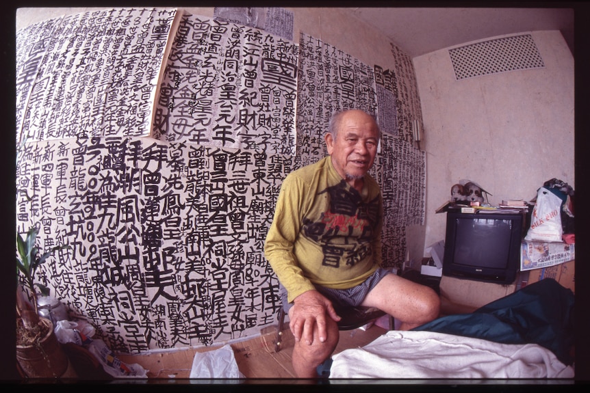A fish eye view of an older Hong Kong man seated in small apartment, behind him is a black ink calligraphy decorated wall.