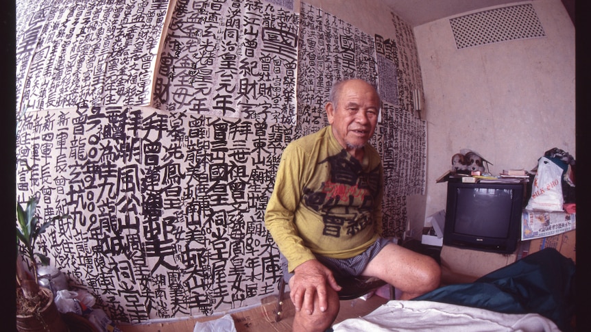 A fish eye view of an older Hong Kong man seated in small apartment, behind him is a black ink calligraphy decorated wall.
