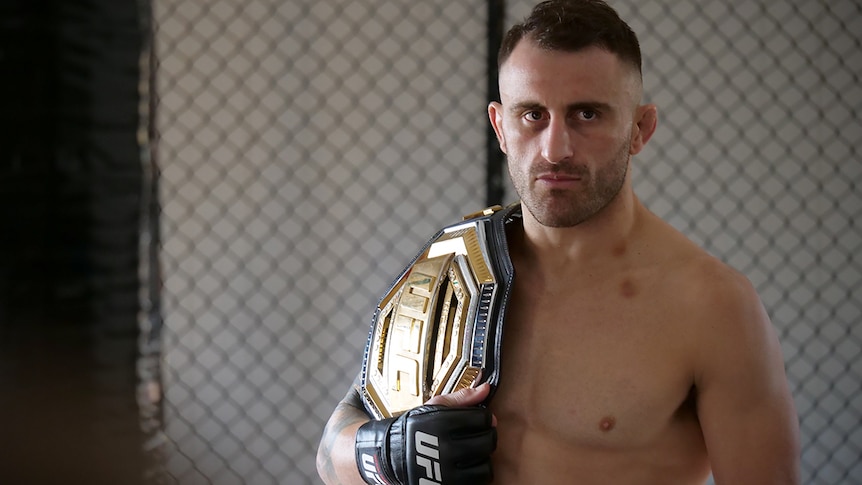 Alex Volkanovski stands shirtless with a belt over his shoulder inside a fighting gym.