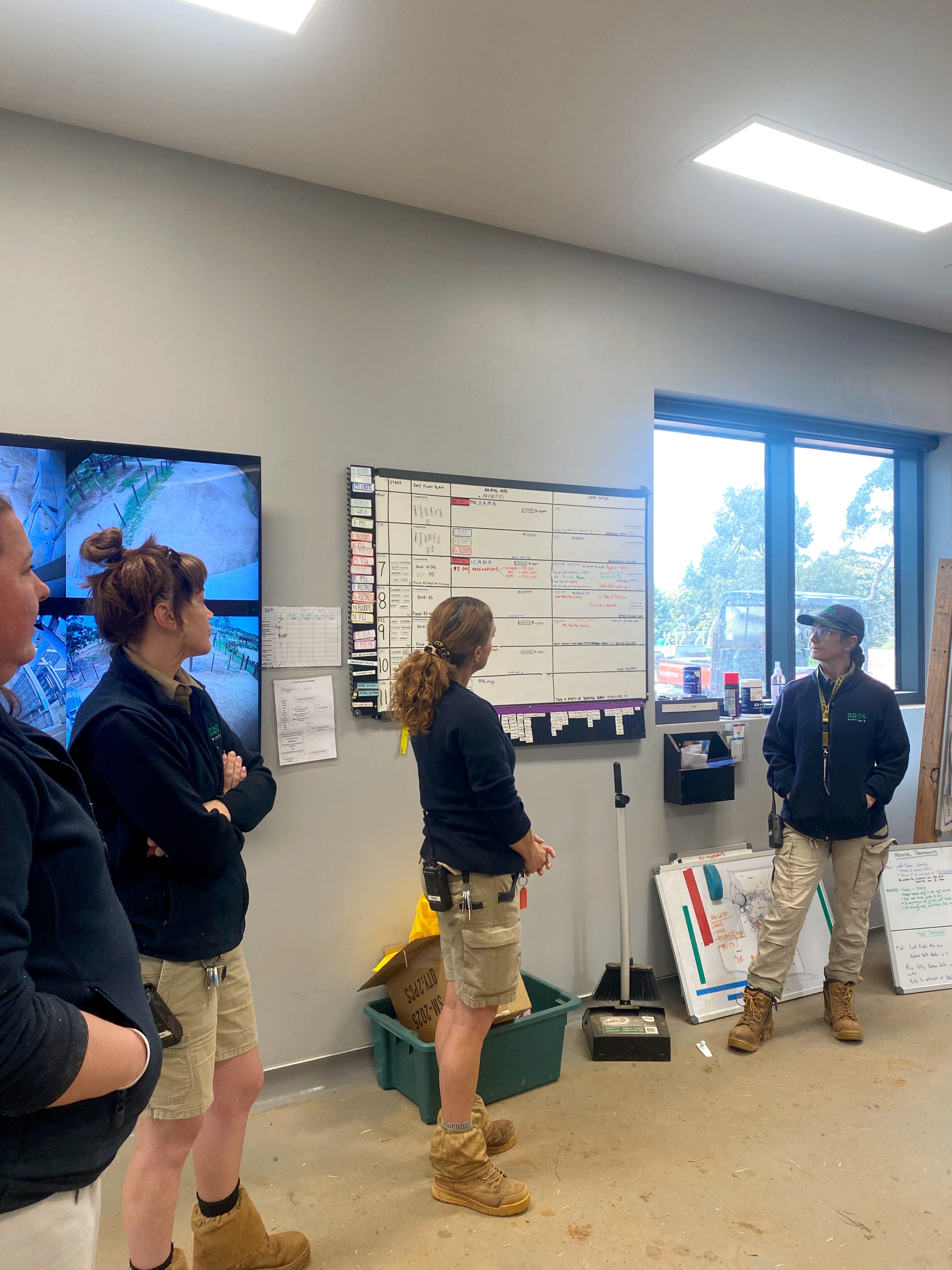 Four people in beige pants and navy jackets standing in a crib room by a whiteboard and CCTV screen.
