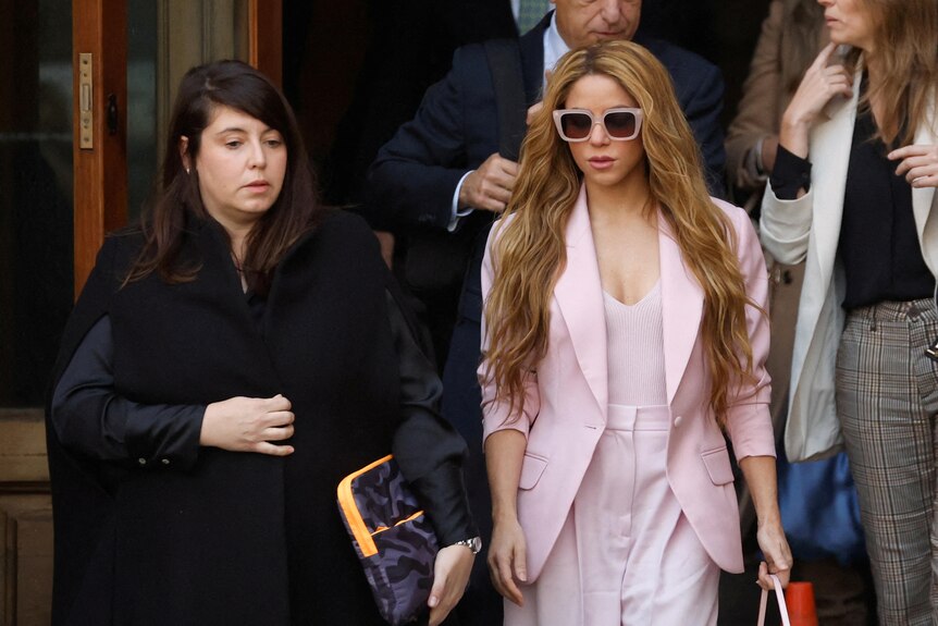 Shakira wearing a pink suit and large sunglasses standing next to a a lawyer in robes. 