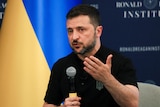 Volodymyr Zelenskyy speaks while holding a microphone in front of a Ukrainian flag.
