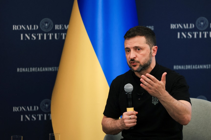 Volodymyr Zelenskyy speaks while holding a microphone in front of a Ukrainian flag.