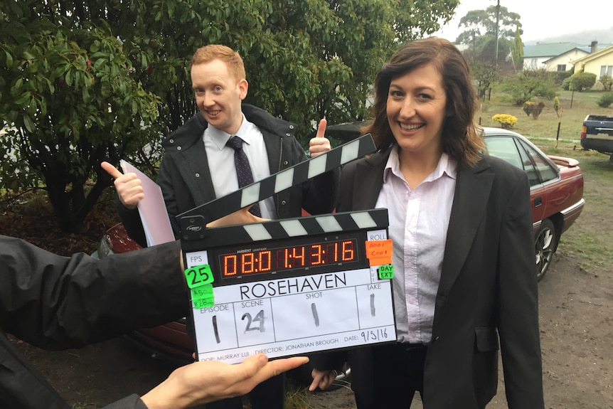 Celia and Luke stand smiling behind a clapperboard for the show Rosehaven