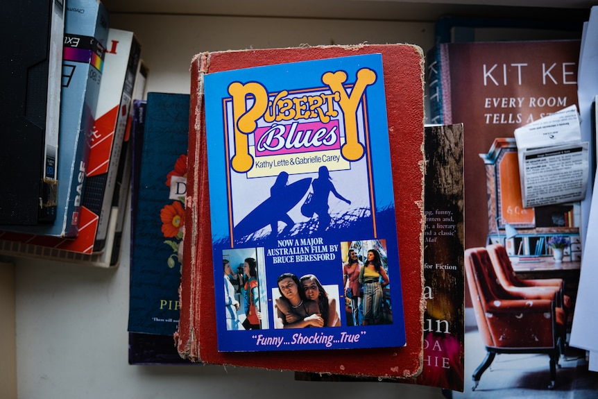 A book sits on a table titled Puberty Blues among a stack of assorted books