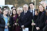 Albanese stands with his hands folded surrounded by a group of mostly women holding flowers.