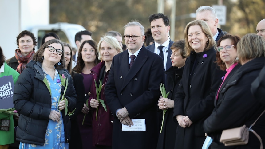 Albanese stands with his hands folded surrounded by a group of mostly women holding flowers.