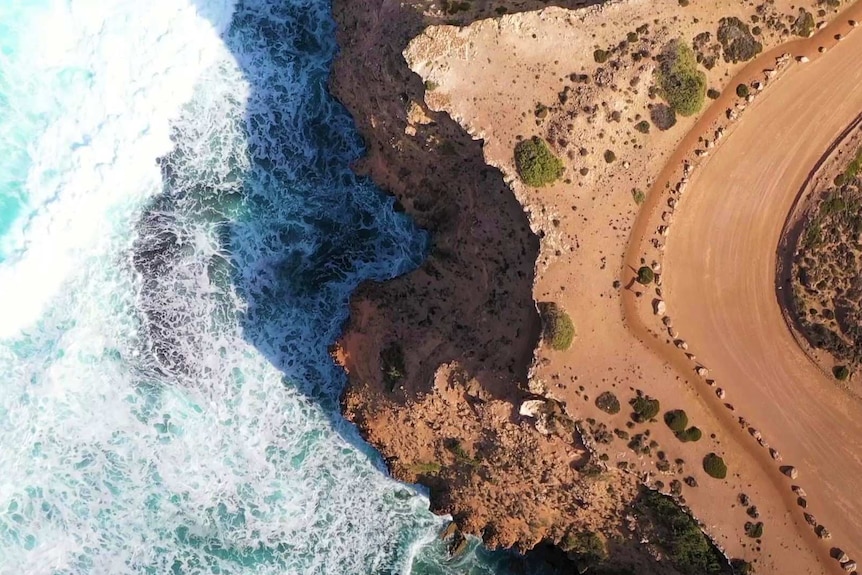 Aerial view of white ocean water on the left crashing against rocky cliff with green vegetation and curved road on right