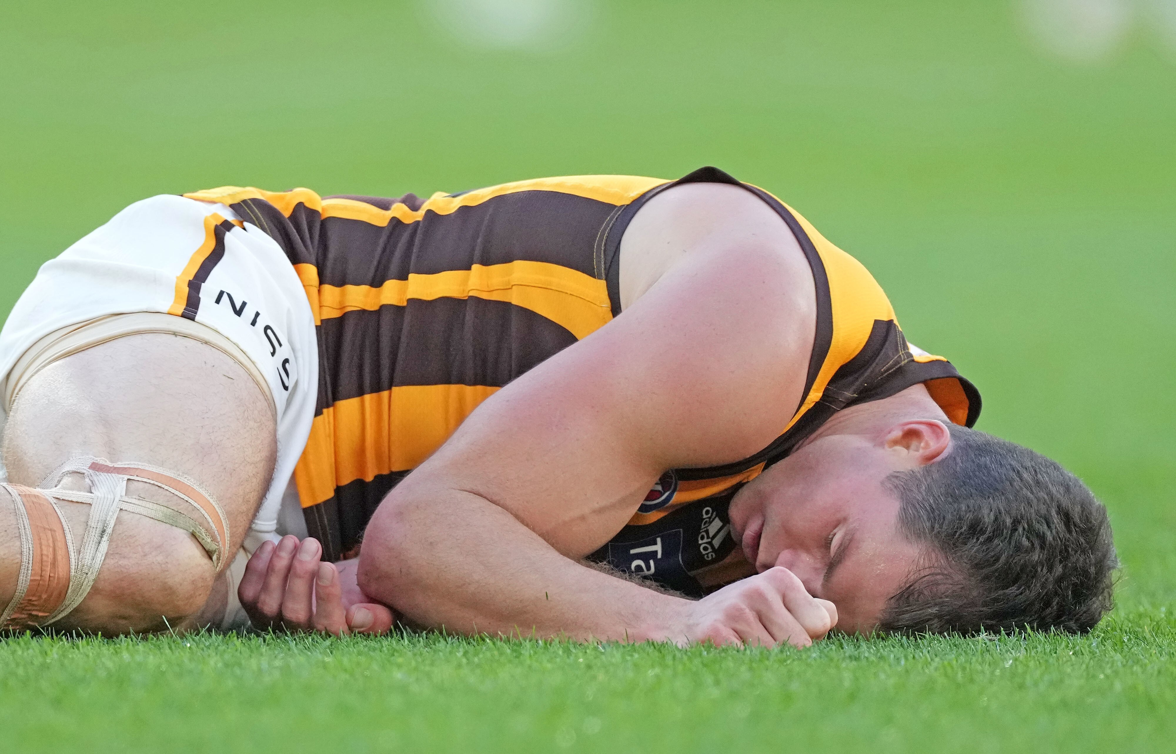 An AFL player lies curled up, motionless, with his eyes closed after an aerial collision during a game. 