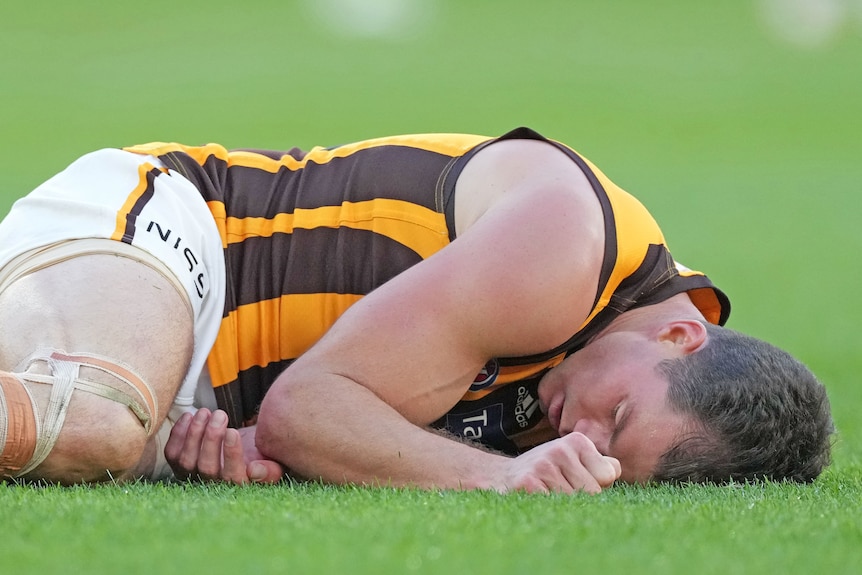 An AFL player lies curled up, motionless, with his eyes closed after an aerial collision during a game. 