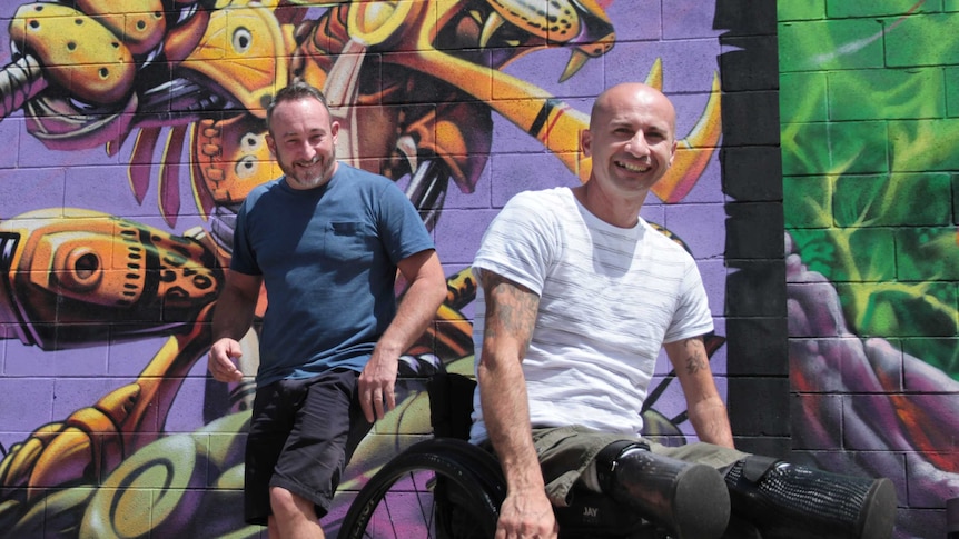 a man in a wheel chair rolling towards camera and man jumping forward from behind him, in front of a purple graffiti wall.