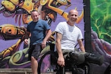 a man in a wheel chair rolling towards camera and man jumping forward from behind him, in front of a purple graffiti wall.