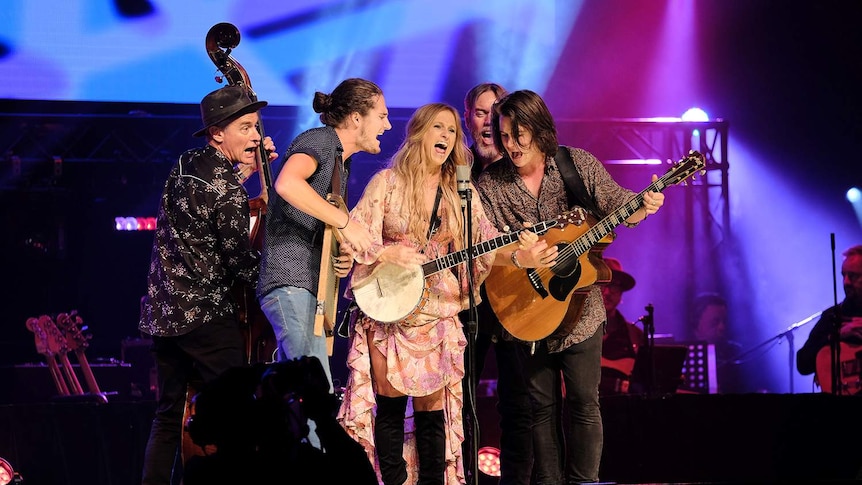 Kasey Chambers and Grizzlee Train clustered around a single microphone on stage, singing.