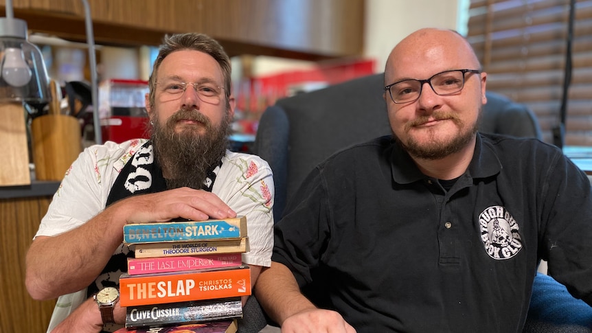 Two men sit side by side holding stacks of books, looking at the camera. 