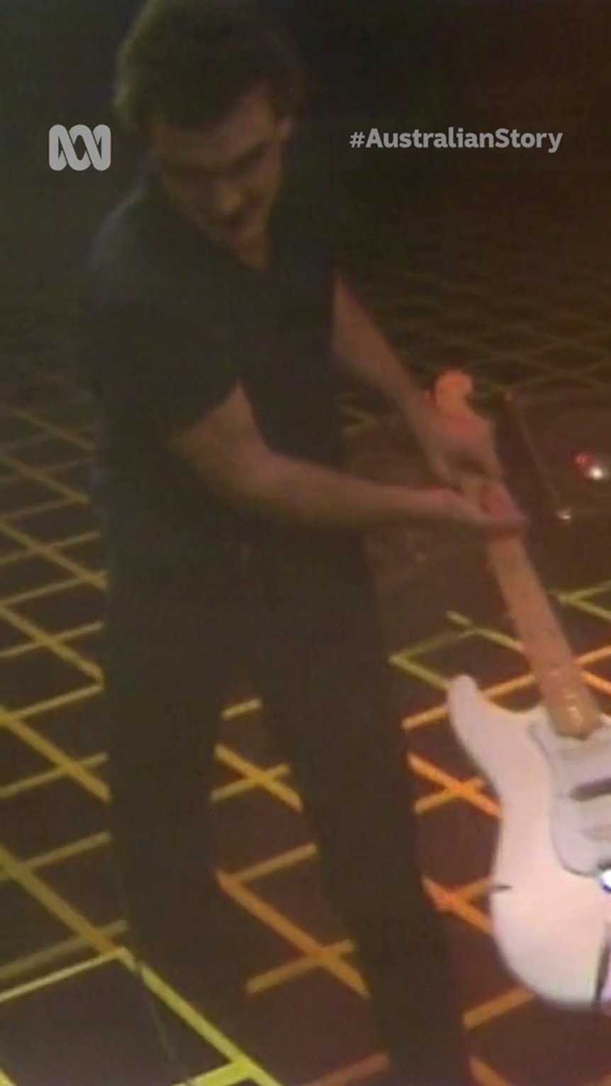 A shot from above shows a man dressed in black with light-tone skin swings a white guitar