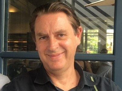 A man with short dark hair and dark clothes sits outside a restaurant, smiling.