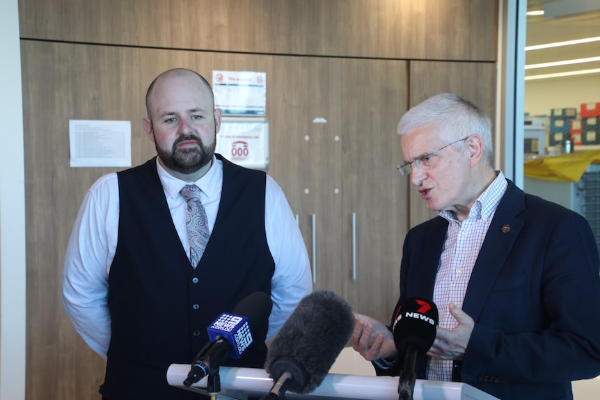 Kieran wears a tie and vest while standing beside Tim who is addressing media