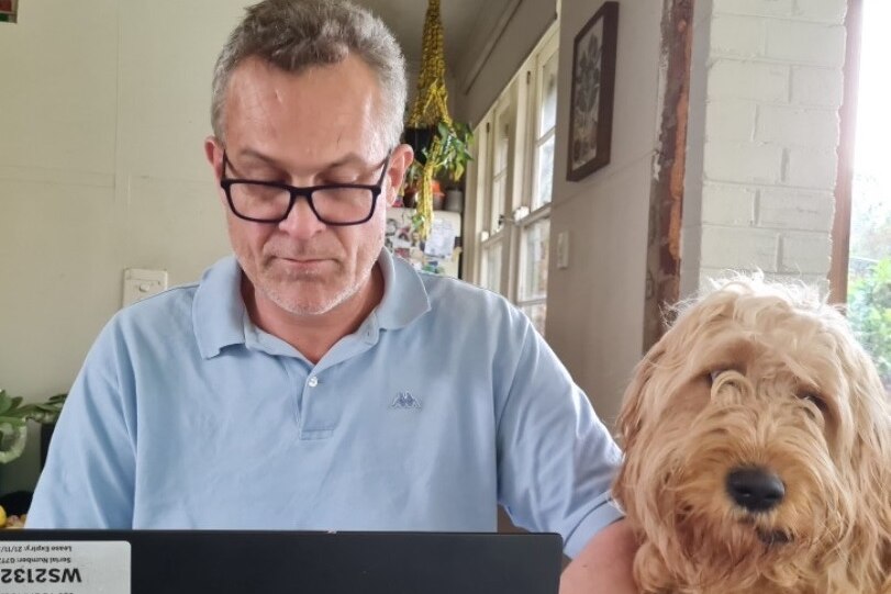 A man wearing glasses sitting at a computer with a dog sitting next to him