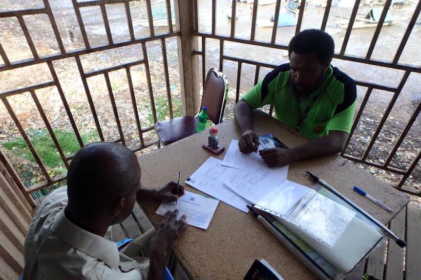 Two Papua New Guinean immigration control officers fill out forms and stamp passports in a secure outside office.
