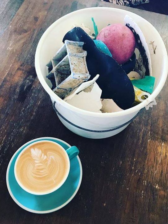A cup of coffee placed next to a bucket of rubbish on a table