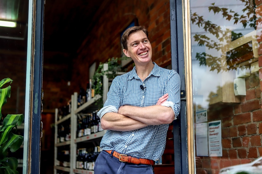 Smiling man with brown hair leans against shop door frame with arms crossed with racks of wine behind