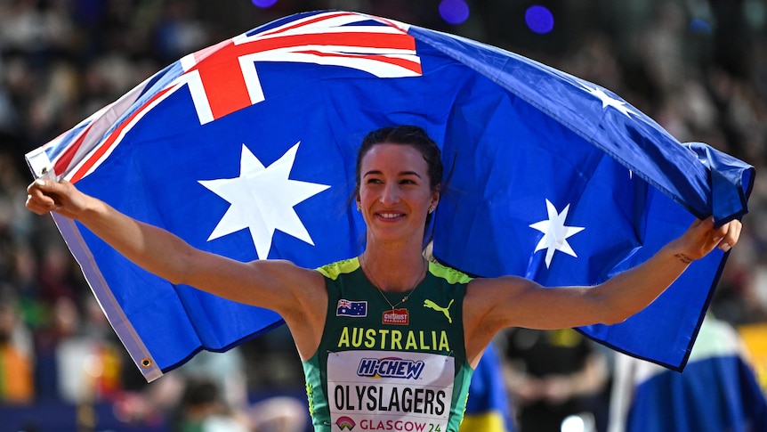 An Australian athlete smiles as she stands with her arms wide holding an Australian flag behind her.