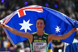 An Australian athlete smiles as she stands with her arms wide holding an Australian flag behind her.