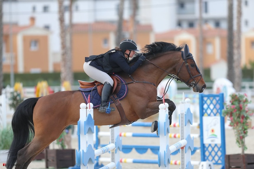 Polina Prigozhina riding on the back of a horse as it jumps over a blue railing.