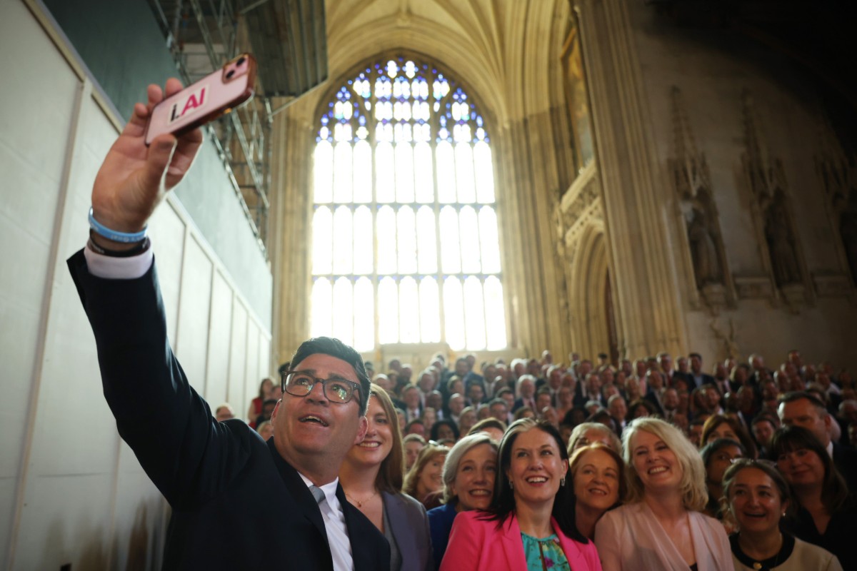 A man in a suit holds a smartphone high in the air, and smiles at it. A large crowd of around 200 people is visible behind him.