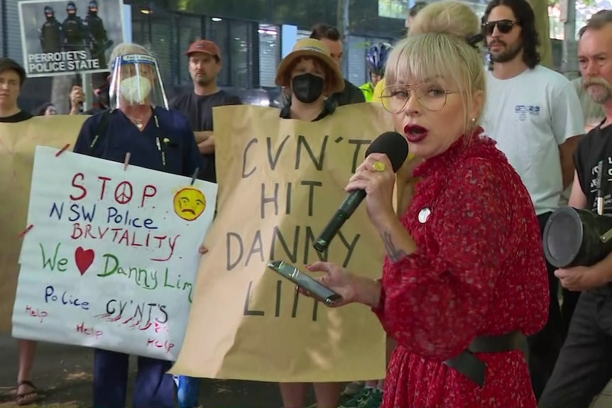 A woman in red speaks into a microphone while protesters with signs stand behind her