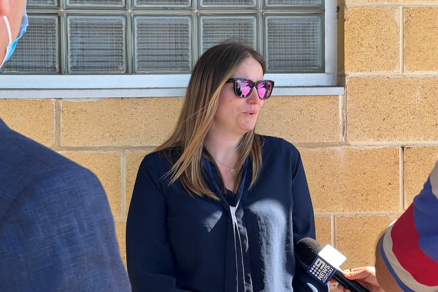 A woman wearing sunglasses speaks into a microphone outside a brick building