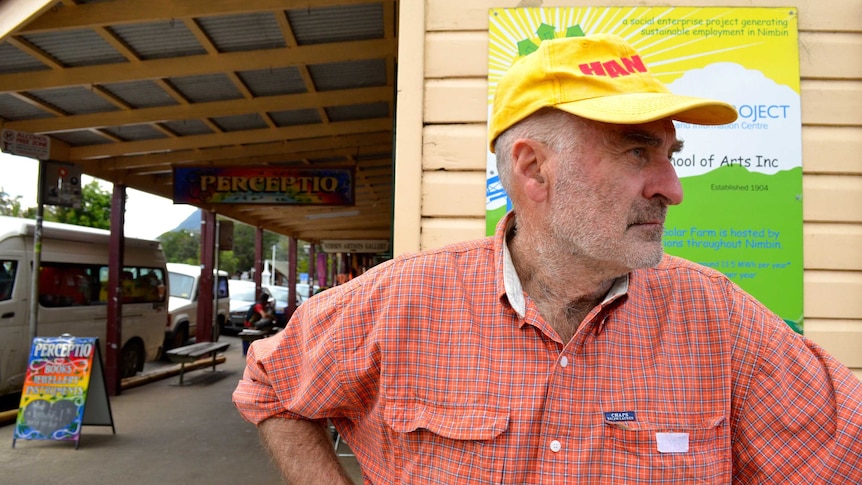 Peter Jackson, stands looking off camera on the main street of Nimbin. Wearing a yellow cap, historic row of Nimbin shops behind