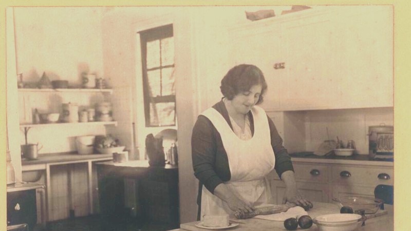 A black and white photo of a woman rolling pastry on a bench in an old fashioned kitchen.