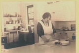 A black and white photo of a woman rolling pastry on a bench in an old fashioned kitchen.