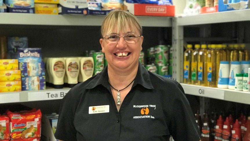 Blonde woman in dark uniform shirt smiles to camera in front of shelves stacked with provisions