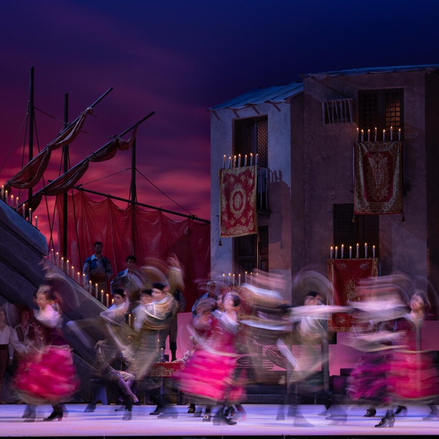 Dancers perform on a stage set depicting a rustic seaside village at sunset with buildings adorned in candles and tapestries.