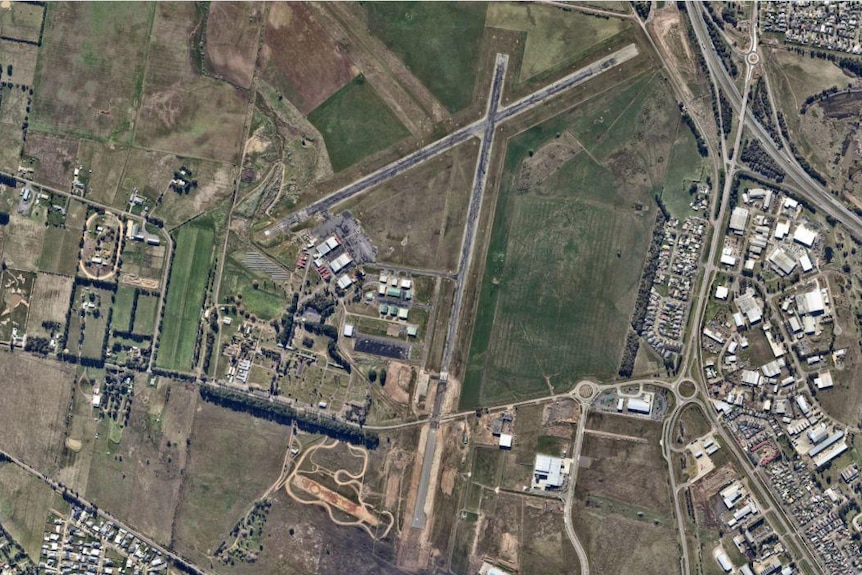 A bird's eye view of an airport showing its runways, construction sites and nearby houses, green paddocks and roads.