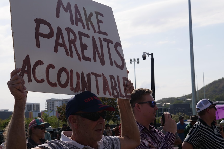 A man holds a sign above his head. It reads make parents accountable. 