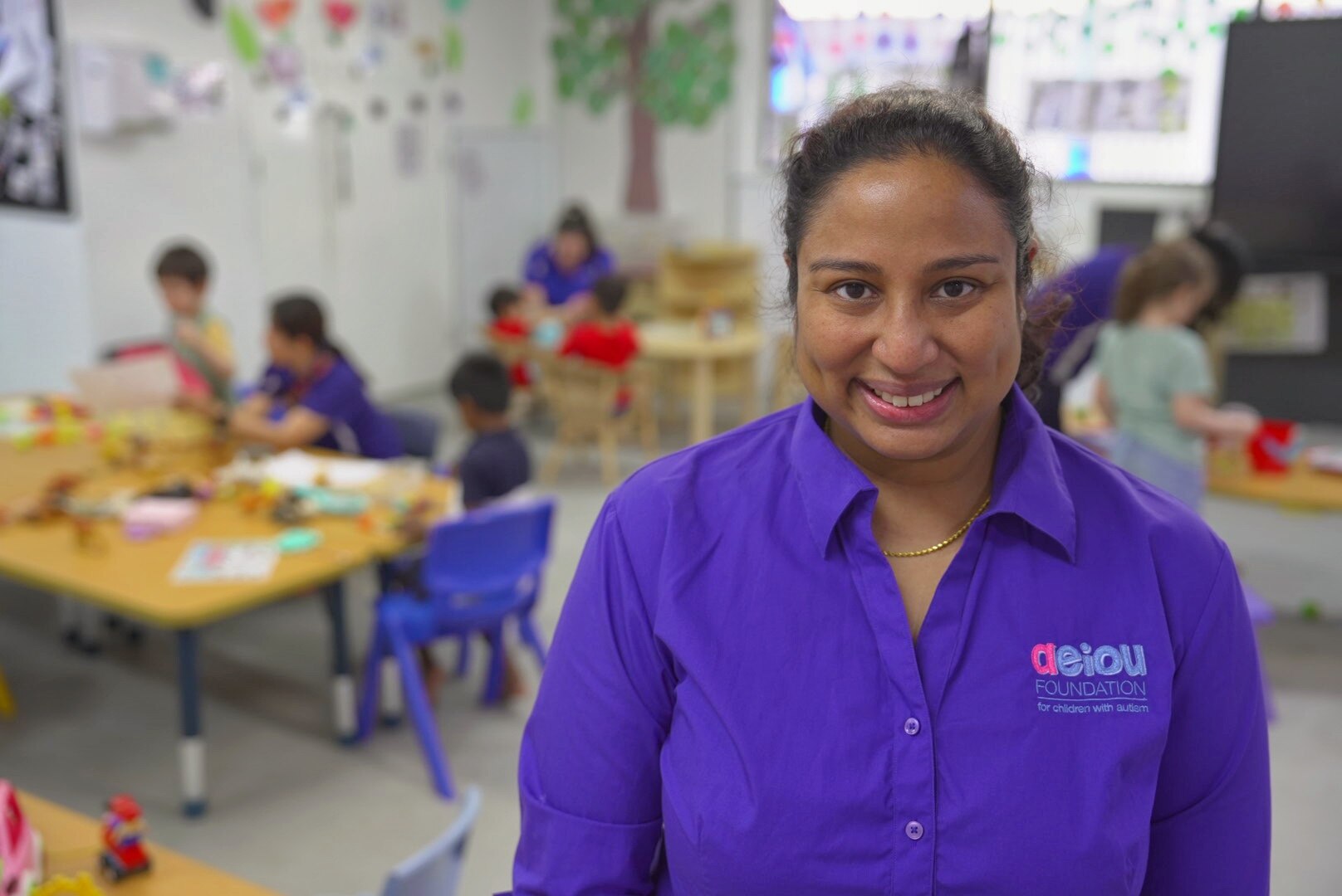 Joanne Stephen wears a purple AEIOU work uniform, children can be seen playing in the background.