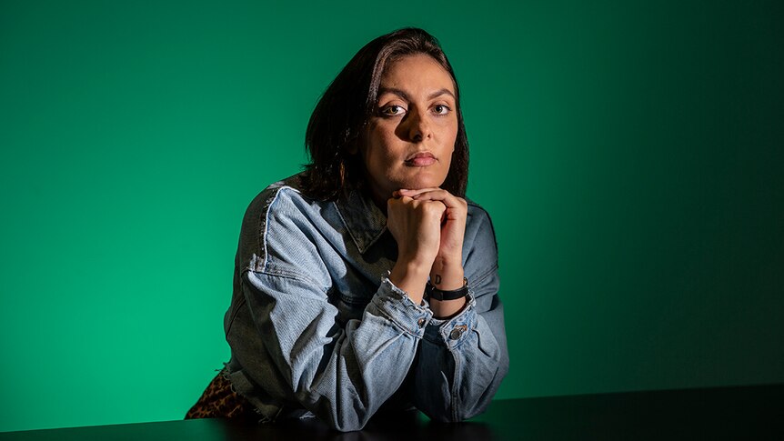 Colour photo of artist Hayley Millar-Baker sitting at a table in front of a green coloured wall.