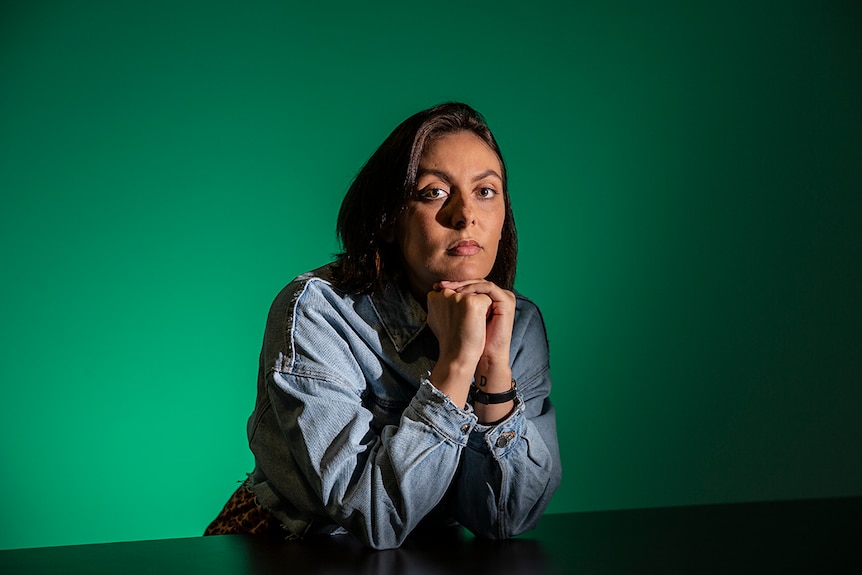 Colour photo of artist Hayley Millar-Baker sitting at a table in front of a green coloured wall.