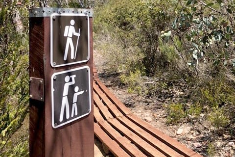 A signpost for walkers and hikers along a boardwalk bush trail with a small mountain the background.